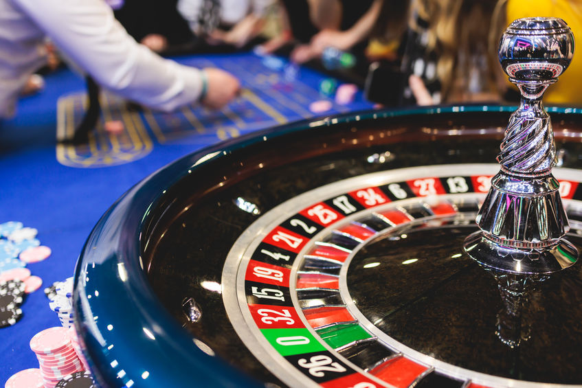 Vibrant casino table with roulette in motion, with casino chips, tokens, the hand of croupier, dollar money and a group of gambling rich people playing bet, blue poker table and deck of cards